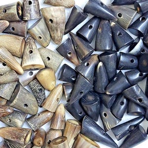 Puede incluir: Una colección de cierres de cuerno en varios tonos de marrón y negro. Cada cierre tiene forma cónica con un agujero perforado en el centro. Los cierres varían en tamaño y textura, algunos muestran patrones de grano natural.