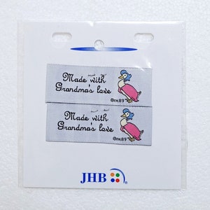 May include: Two white fabric labels with a pink and blue duck illustration and the text "Made with Grandma's love" and the copyright symbol followed by "FW.89". The labels are packaged in a clear plastic bag with the text "JHB" and a logo of four coloured dots.