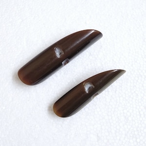 May include: Two dark brown toggle buttons with a glossy finish. Each button has a rounded end and a rectangular hole for sewing. The buttons are made of a smooth material and are ideal for coats or jackets.
