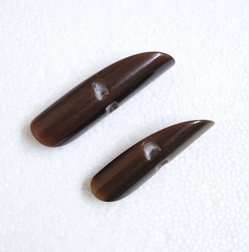 May include: Two dark brown toggle buttons with a glossy finish. Each button has a rounded end and a rectangular hole for sewing. The buttons are made of a smooth material and are ideal for coats or jackets.