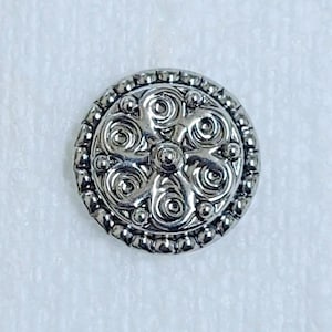 May include: A silver-colored button with an ornate, swirling design. The button has a central flower-like pattern surrounded by six spiral shapes. The outer edge is detailed with small, raised beads.