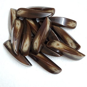May include: A collection of brown, elongated beads with a glossy finish. Each bead has a lighter, cream-colored pattern running through its center, creating a natural, organic look. The beads are scattered on a white surface.