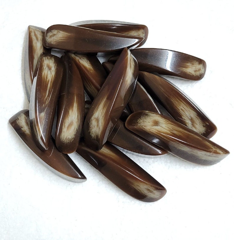 May include: A collection of brown, elongated beads with a glossy finish. Each bead has a lighter, cream-colored pattern running through its center, creating a natural, organic look. The beads are scattered on a white surface.