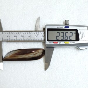 May include: A polished, elongated gemstone with a mix of brown and cream tones, measured at 2.362 inches using a digital caliper. The stone has a smooth, curved shape and a glossy finish, set against a white background.