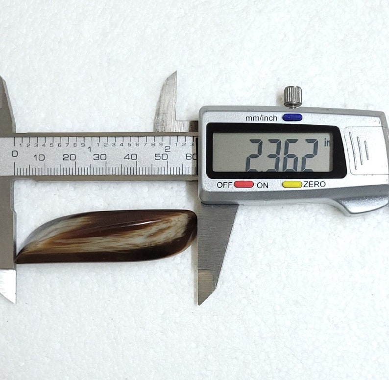 May include: A polished, elongated gemstone with a mix of brown and cream tones, measured at 2.362 inches using a digital caliper. The stone has a smooth, curved shape and a glossy finish, set against a white background.
