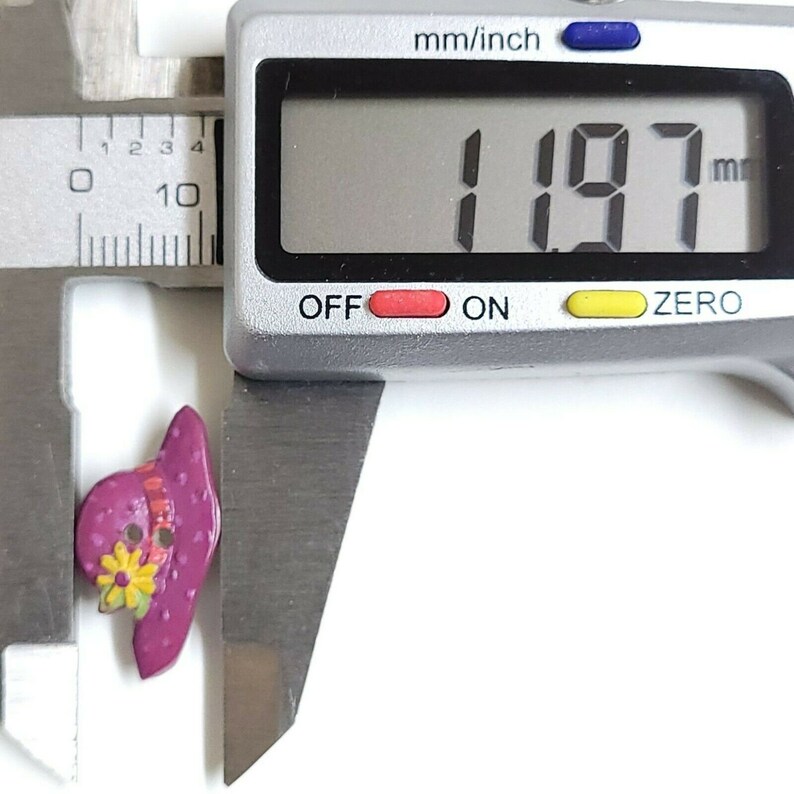 May include: A close-up of a purple and yellow button shaped like a hat with a flower design. The button is being measured with a caliper, which reads 1.97 mm.
