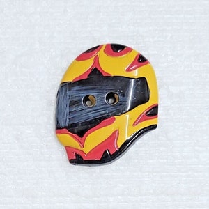 Könnte beinhalten: Ein gelber, roter und schwarzer Keramikknopf in Form eines Motorradhelms. Der Helm hat ein schwarzes Visier mit zwei Löchern zum Nähen.