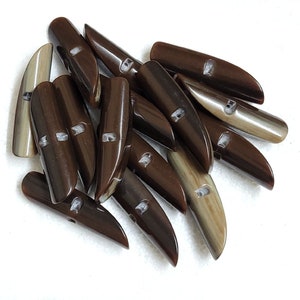 May include: A collection of brown and tan toggle buttons. The buttons are elongated with a rounded top and a pointed end. Each button has two holes for sewing. The buttons are made of a glossy material.