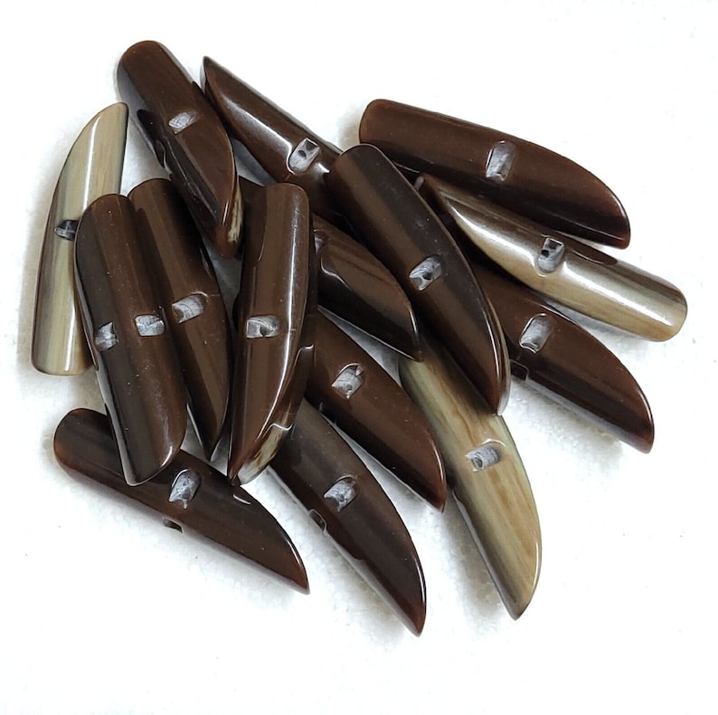 May include: A collection of brown and tan toggle buttons. The buttons are elongated with a rounded top and a pointed end. Each button has two holes for sewing. The buttons are made of a glossy material.