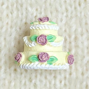 Puede incluir: Una figura de pastel de tres niveles en color amarillo pálido decorada con bordes de glaseado blanco y pequeñas rosas moradas con hojas verdes. El pastel en miniatura tiene un acabado liso y adornos florales en cada uno de los niveles.
