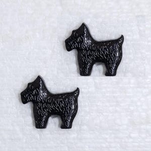 May include: Two black dog-shaped buttons. The buttons are glossy and have a textured surface, with details suggesting fur. The buttons are positioned on a white, textured background.