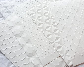 Könnte beinhalten: Sechs weiße, strukturierte Papierbögen mit verschiedenen Prägemustern. Die Muster umfassen eine Wabe, eine geometrische Form, ein Punktmuster, ein Sternmuster und zwei verschiedene geometrische Muster.