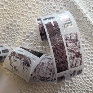 Könnte beinhalten: Dekorative Washi-Tape-Rollen mit verschiedenen Designs. Eine Rolle zeigt ein Frauengesicht, eine andere eine Stadtlandschaft mit Glitzer und eine dritte ein geometrisches Muster. Das Tape ist weiß mit schwarzen, braunen und silbernen Details.