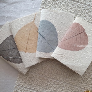 Può includere: Quattro piccoli quaderni quadrati con copertine bianche strutturate, ognuno con una foglia pressata in diversi colori: grigio, beige, blu e rosa. Le foglie sono centrate sulle copertine. I quaderni sono disposti su una superficie di pizzo bianco.