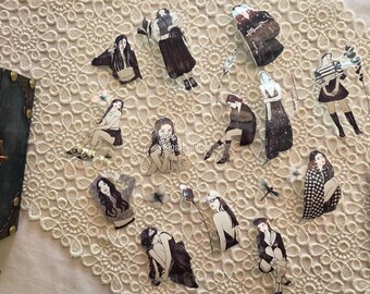 May include: A collection of decorative stickers featuring illustrations of women in various poses and outfits. The stickers are in black and white, with some accents of color. The stickers are arranged on a cream-colored, lace-like surface.