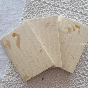 Könnte beinhalten: Drei kleine, cremefarbene Notizbücher mit einem strukturierten, handgefertigten Papieraussehen. Jedes Notizbuch hat ein einzigartiges Design mit goldfarbenen Akzenten und sichtbaren Papierfasern. Die Notizbücher sind auf einer weißen Spitzentischdecke angeordnet.