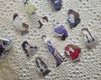 May include: A collection of illustrated stickers featuring various women in different poses and outfits. The stickers are placed on a white, textured surface. A roll of decorative tape is visible in the corner.