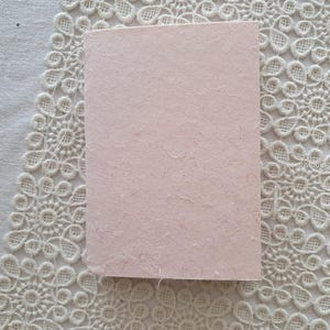 May include: A rectangular, pale pink card with a textured surface. The card is set against a white, floral lace background. The card appears to be made of handmade paper.