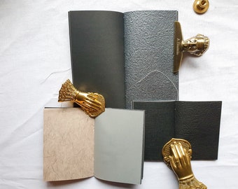 May include: Four small notebooks with different covers, including black, gray, and beige. Each notebook is closed and has a brass paperclip attached. The paperclips are in the shape of a hand.