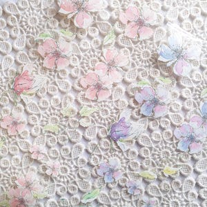 Könnte beinhalten: Ein weißer Spitzenstoff-Hintergrund mit zarten Aquarell-Blumenmustern in Rosa-, Blau- und Lilatönen.