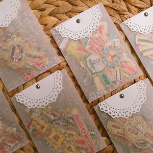 Op de afbeelding: Zes transparante plastic enveloppen met witte kantversiering en een zilveren knoopsluiting. Elke envelop bevat een verzameling vintage sigarenlabels.