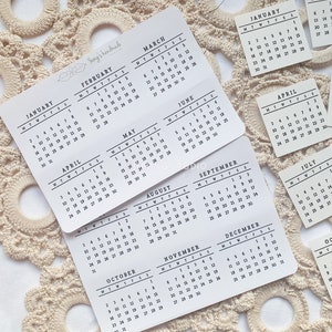 Könnte beinhalten: Ein Satz von zwölf kleinen weißen Karten mit schwarzem Text, jede mit einem anderen Monat des Jahres im Kalenderformat. Die Karten sind in einem Raster auf einer weißen Oberfläche mit einer gehäkelten Unterlage angeordnet.