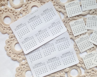 May include: A set of twelve small white cards with black text, each featuring a different month of the year in a calendar format. The cards are arranged in a grid pattern on a white surface with a crocheted doily underneath.