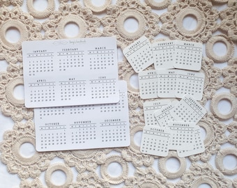 May include: A set of 12 small white cards with black text, each featuring a different month of the year in a calendar format. The cards are arranged on a white crocheted surface.