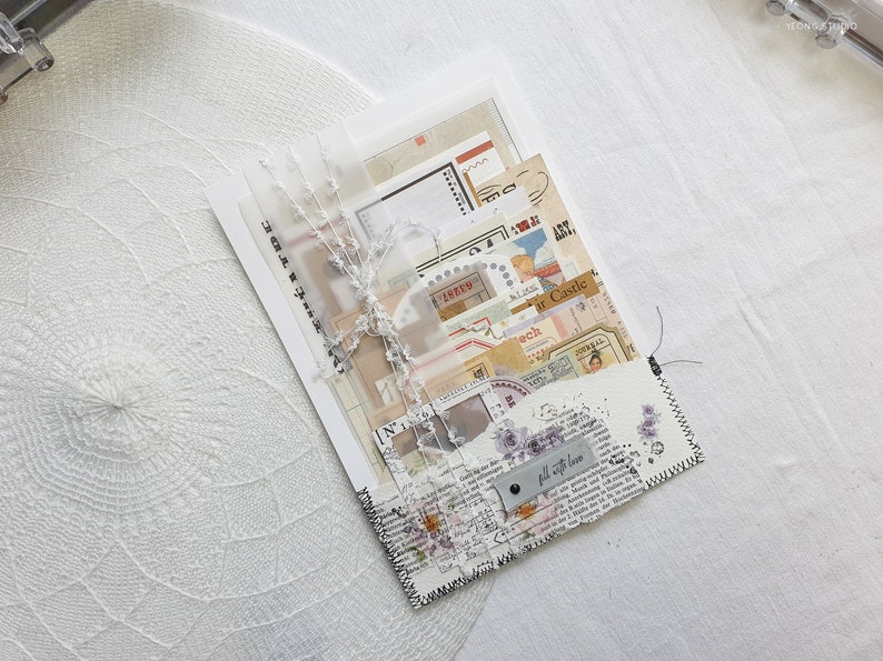 K&ouml;nnte beinhalten: Ein wei&szlig;er Umschlag mit einem gen&auml;hten Rand, gef&uuml;llt mit Papier-Ephemera im Vintage-Stil, darunter Dekorpapier, Aufkleber und ein Etikett mit der Aufschrift "Mit Liebe f&uuml;llen".