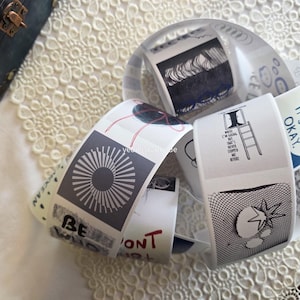 Könnte beinhalten: Eine Sammlung weißer Papieretiketten mit verschiedenen schwarzen und grauen grafischen Designs und Texten, darunter ein Sonnenstrahl, eine Leiter und die Worte „BE WHO YOU WANT“. Die Etiketten sind gerollt und kreisförmig angeordnet.