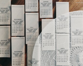 Könnte beinhalten: Ein Satz von zwölf weißen Karten mit schwarzem Text, jede mit einem anderen Monat des Jahres in deutscher Sprache. Die Karten liegen auf einer weißen Häkelunterlage.