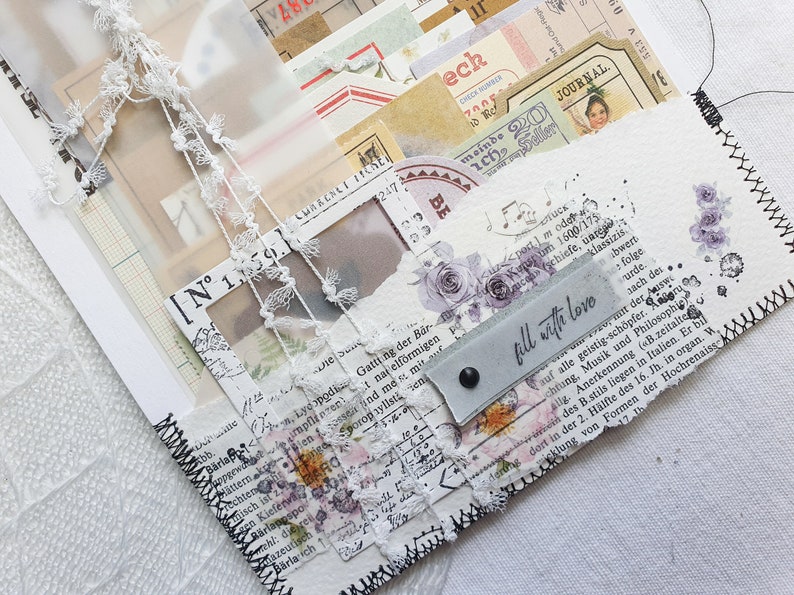 K&ouml;nnte beinhalten: Ein wei&szlig;es Papierjournal mit einem schwarzen, gesteppten Rand. Das Journal ist mit Spitze, Papierschnipseln und einem Etikett verziert, auf dem "f&uuml;lle mit Liebe" steht.