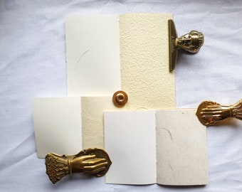 May include: Four blank paper notebooks with decorative brass clips. Two clips feature a hand design, and one features a bust of a man. The notebooks are arranged in a square shape on a white surface.