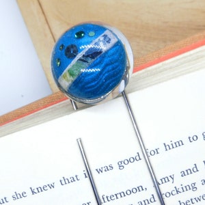 Könnte beinhalten: Ein dekorativer Lesezeichen mit einem runden, blau-weißen Design und silberfarbenem Metall. Das Lesezeichen wird in ein Buch eingelegt und verleiht dem Lesen einen Hauch von Stil. Das Design umfasst blaue Pailletten und eine Landschaft.