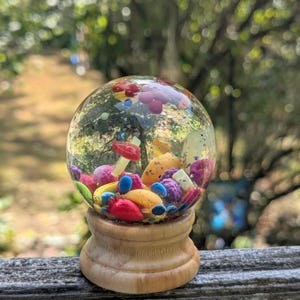 May include: A clear glass snow globe with colorful miniature mushrooms, berries, and confetti inside. The globe sits on a wooden base. The scene inside includes red, yellow, blue, and purple elements, creating a whimsical, decorative item.