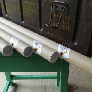 K&ouml;nnte beinhalten: Mehrere aufgerollte Poster in Pappr&ouml;hren, mit wei&szlig;en Endkappen und Etiketten. Die R&ouml;hren liegen auf einer gr&uuml;nen Oberfl&auml;che, im Hintergrund eine dunkle Metallbox. Die Etiketten tragen Text und ein Logo.