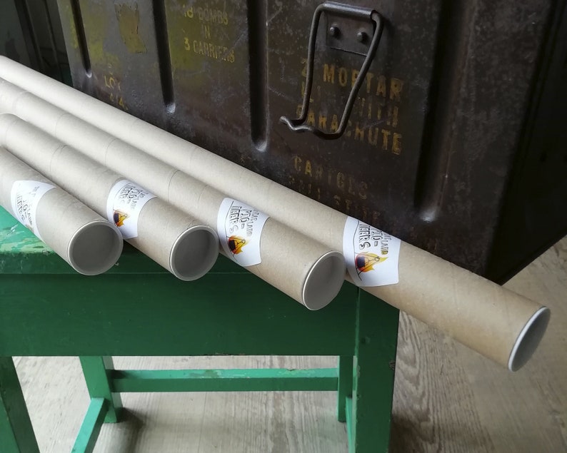 K&ouml;nnte beinhalten: Mehrere aufgerollte Poster in Pappr&ouml;hren, mit wei&szlig;en Endkappen und Etiketten. Die R&ouml;hren liegen auf einer gr&uuml;nen Oberfl&auml;che, im Hintergrund eine dunkle Metallbox. Die Etiketten tragen Text und ein Logo.
