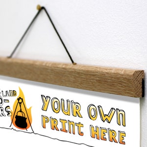 K&ouml;nnte beinhalten: Ein h&ouml;lzerner Posterhalter mit schwarzem Kordel, der einen wei&szlig;en Druck h&auml;lt. Der Druck zeigt den Text "THE FLAT LAND PIONEERS" und "YOUR OWN PRINT HERE" in einer verspielten Schriftart, zusammen mit einer Lagerfeuer-Illustration.