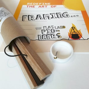K&ouml;nnte beinhalten: Ein Poster-Rahmen-Set mit Holzleisten, einer schwarzen Kordel und einem wei&szlig;en Kunststoffteil mit einem Metallstift. Das Set ist in einer braunen Pappr&ouml;hre verpackt. Das Poster im Hintergrund tr&auml;gt den Text "Redefine the Art of Framing..." und eine Campingszene.