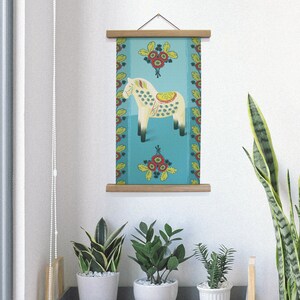 K&ouml;nnte beinhalten: Ein gerahmter Wandkunstdruck mit einem stilisierten wei&szlig;en Pferd mit gelbem Sattel und gr&uuml;nen Punkten auf t&uuml;rkisfarbenem Hintergrund. Der Druck ist mit floralen Mustern in Rot, Gelb und Gr&uuml;n verziert und wird mit einem Holzrahmen pr&auml;sentiert.
