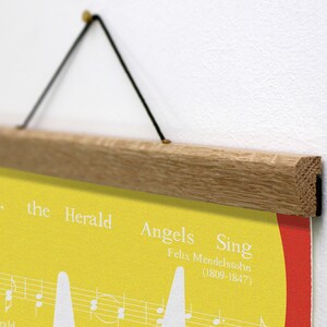K&ouml;nnte beinhalten: Ein Poster mit gelbem Hintergrund und wei&szlig;em Text, einschlie&szlig;lich Noten und den Worten "Hark, the Herald Angels Sing". Das Poster wird von einem hellbraunen Holzrahmen und schwarzem Faden gehalten.