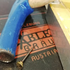 K&ouml;nnte beinhalten: Ein Werkzeug mit blauem Griff und Holzende, das auf einer dunklen Oberfl&auml;che mit dem Text "SALZBURG" und "AUSTRIA" liegt. Ein kleines Metallwerkzeug mit dekorativem Kopf ist ebenfalls vorhanden.
