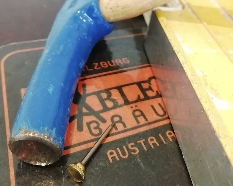 K&ouml;nnte beinhalten: Ein Werkzeug mit blauem Griff und Holzende, das auf einer dunklen Oberfl&auml;che mit dem Text "SALZBURG" und "AUSTRIA" liegt. Ein kleines Metallwerkzeug mit dekorativem Kopf ist ebenfalls vorhanden.
