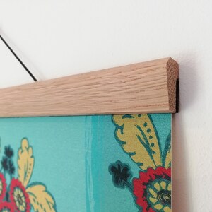 K&ouml;nnte beinhalten: Ein gerahmter Druck mit einem hellen Holzrahmen und einer schwarzen Aufh&auml;ngeschnur. Der Druck zeigt einen t&uuml;rkisfarbenen Hintergrund mit floralen Mustern in Rot, Gelb und Dunkelblau. Der Rahmen besteht aus hellem Holz.