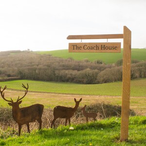 May include: A wooden post sign hanging a rectangular wooden plaque with the text The Coach House in black lettering, accompanied by three dark brown metal deer silhouettes of varying sizes standing in a row on the grass.