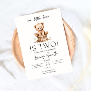 May include: A white invitation card with a brown bear sitting on a tree stump. The text on the card reads "our little bear is two!" and includes the name "Henry Smith", the date "October 24, 2024", and the time "at 4 pm".