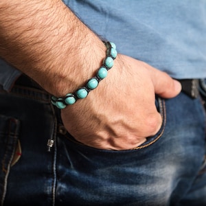 Puede incluir: Un brazalete de cuero negro con cuentas turquesas. El brazalete se lleva en la muñeca.