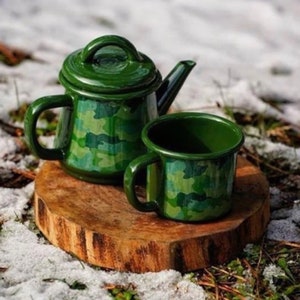 Puede incluir: Un juego de tetera y taza de esmalte verde camuflaje sobre una superficie de madera. La tetera tiene una tapa y un asa. La taza tiene un asa.
