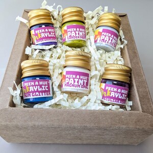 Close up image of a cheerful kraft gift box filled with six petite glass jars of sparkly iridescent paint. Each jar has a different color and a colorful label on it, and a matte metallic twist off gold lid. The box is filled with recycled off-white crinkle paper.