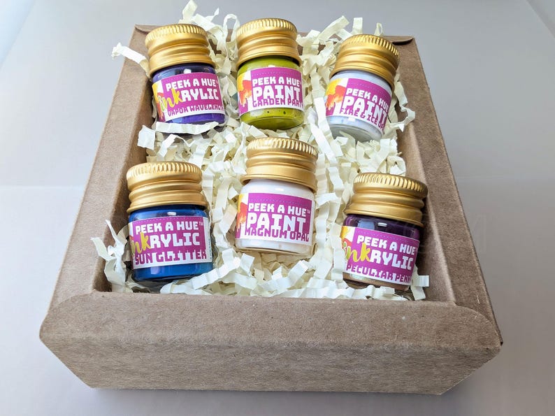 Close up image of a cheerful kraft gift box filled with six petite glass jars of sparkly iridescent paint. Each jar has a different color and a colorful label on it, and a matte metallic twist off gold lid. The box is filled with recycled off-white crinkle paper.